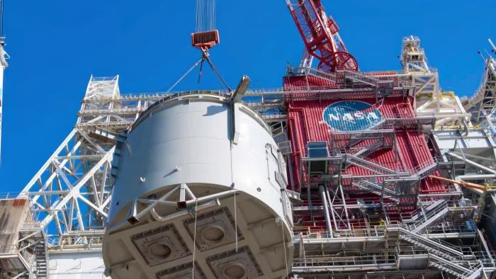 NASA hoisting a huge cylindrical rocket section stories high next to a large testing center.