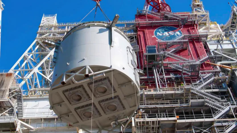 NASA hoisting a huge cylindrical rocket section stories high next to a large testing center.