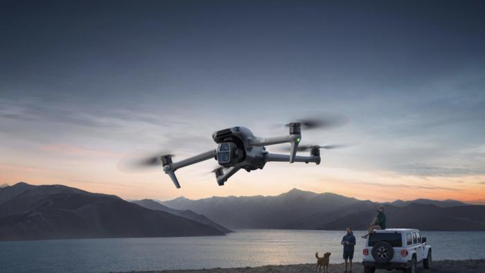 A small flying machine against the sky with two humans, a dog and a vehicle in the background.